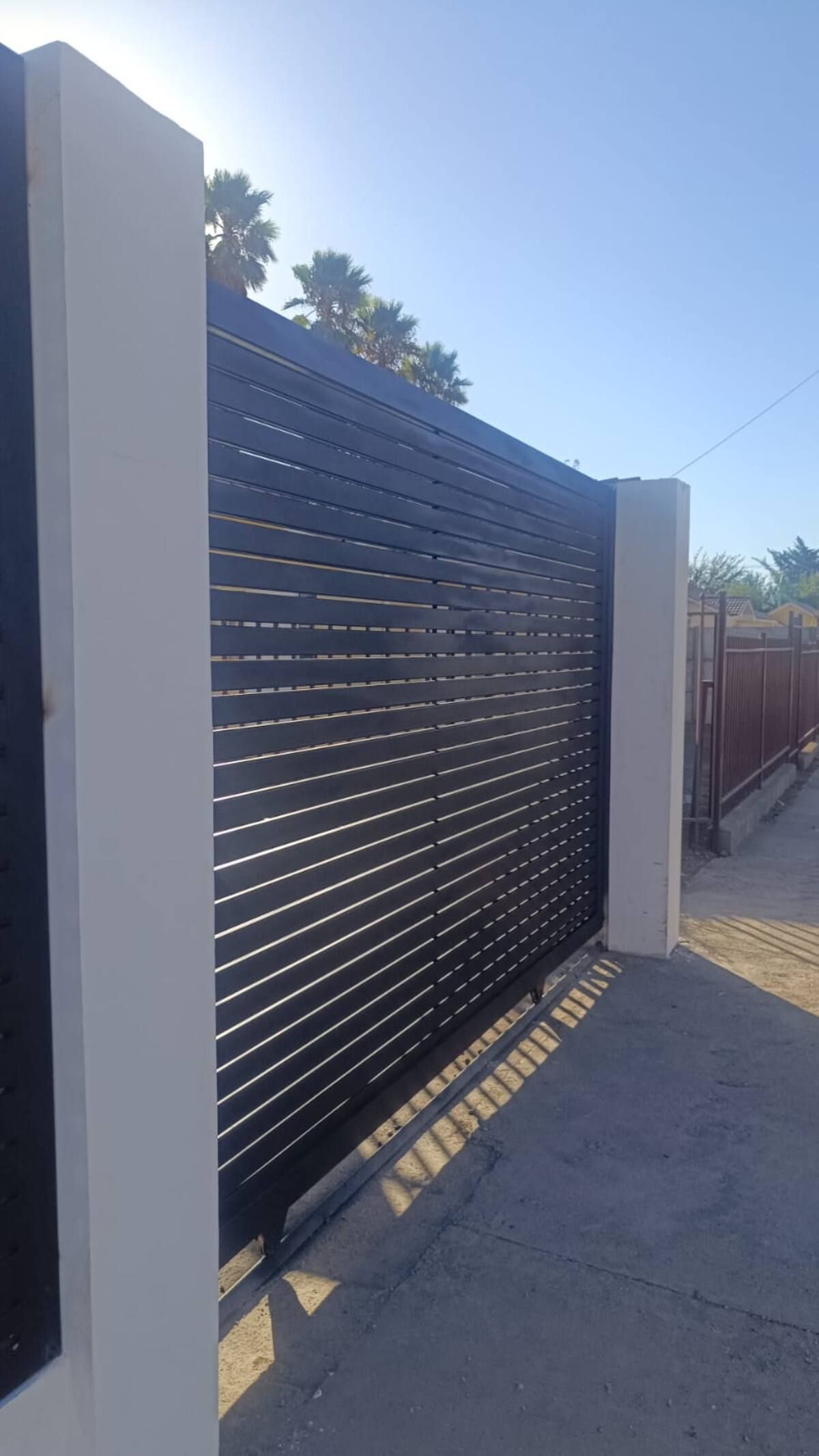 Una reja de metal negro con listones horizontales, pilares blancos y suelo empedrado.