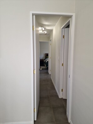 A narrow hallway with white walls and a tiled floor leading to a bedroom with a bed and a dresser.