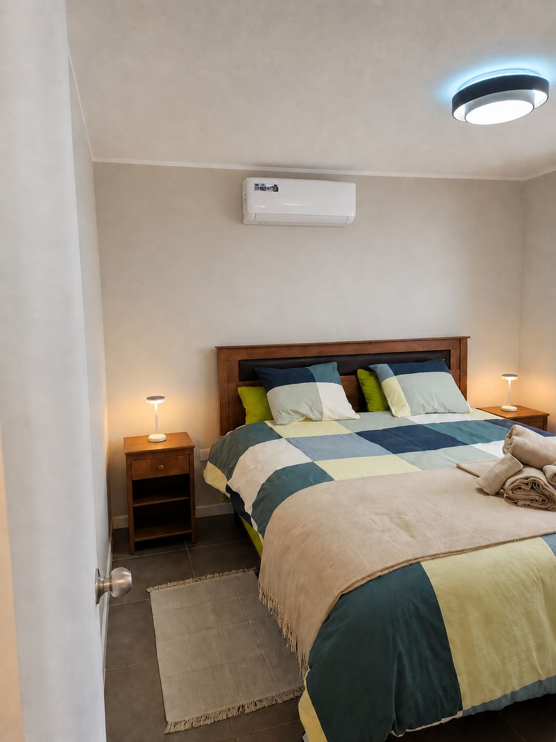 A bedroom with a bed, bedside tables, a wall-mounted air conditioner, and a ceiling light fixture.