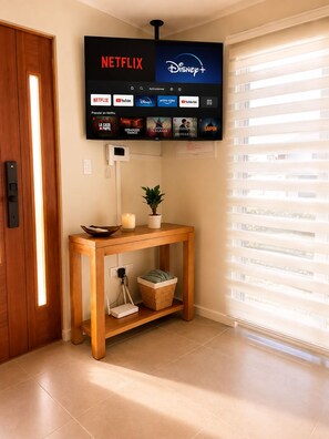 Un televisor de pantalla plana montado en la pared mostrando los logotipos de Netflix y Disney+, una mesa consola de madera con una planta, una vela y una cesta, y una puerta con interruptor de luz.