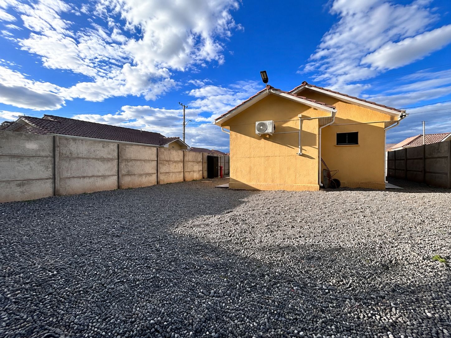 Un petit bâtiment jaune avec une allée de gravier, un mur en béton et un ciel bleu clair parsemé de nuages.