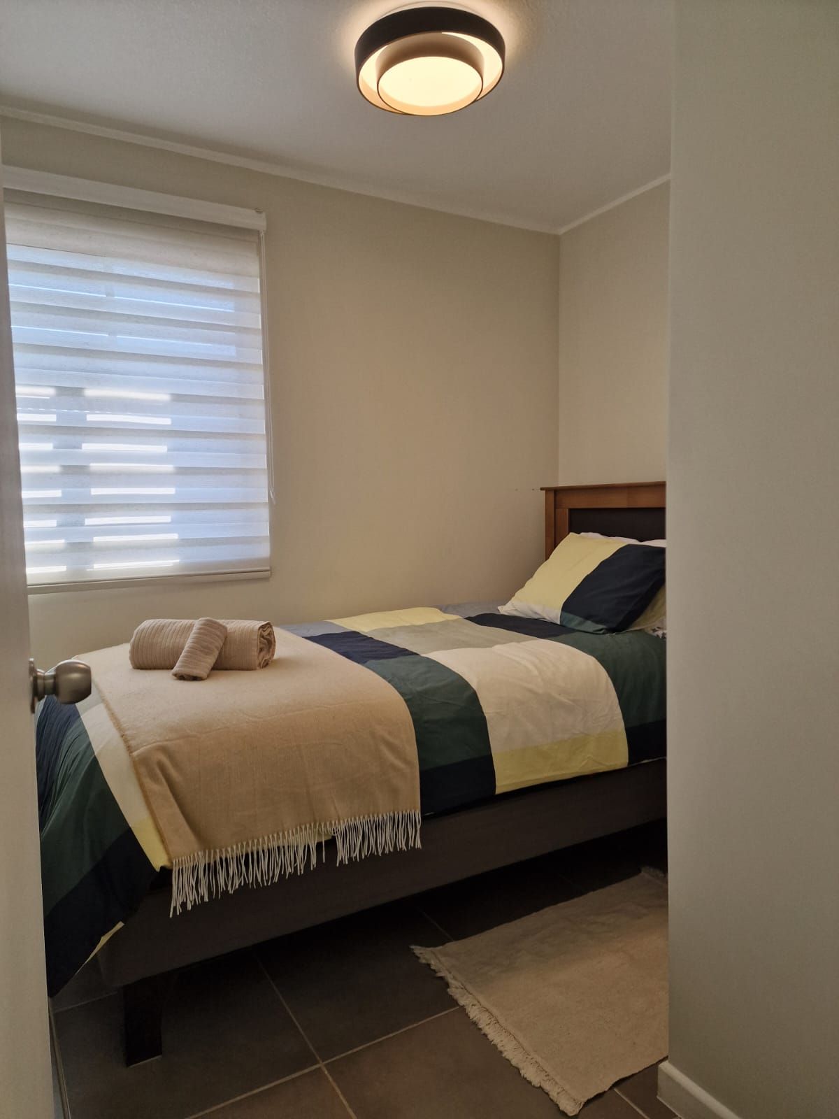 A bedroom with a bed, a window with blinds, a ceiling light, and a door.