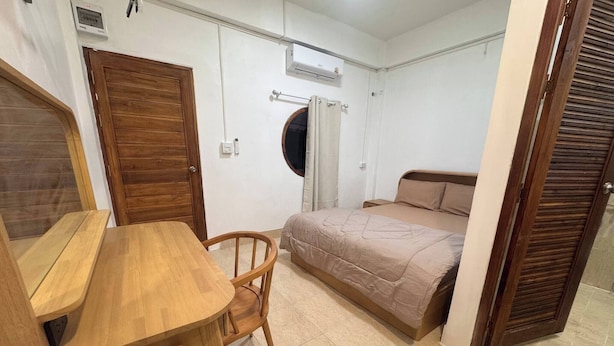 Een kamer met een houten bureau, een stoel, een bed met een beige dekbedovertrek, een raam met gordijnen en een houten deur.