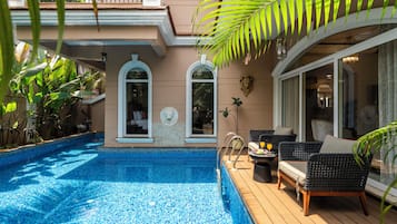 A swimming pool with a tiled deck, surrounded by lush greenery and a covered seating area with wicker furniture.