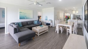 Ein modernes Wohnzimmer mit einem grauen Ecksofa, einem Holztisch und einem Essbereich mit weißem Tisch und Stühlen.