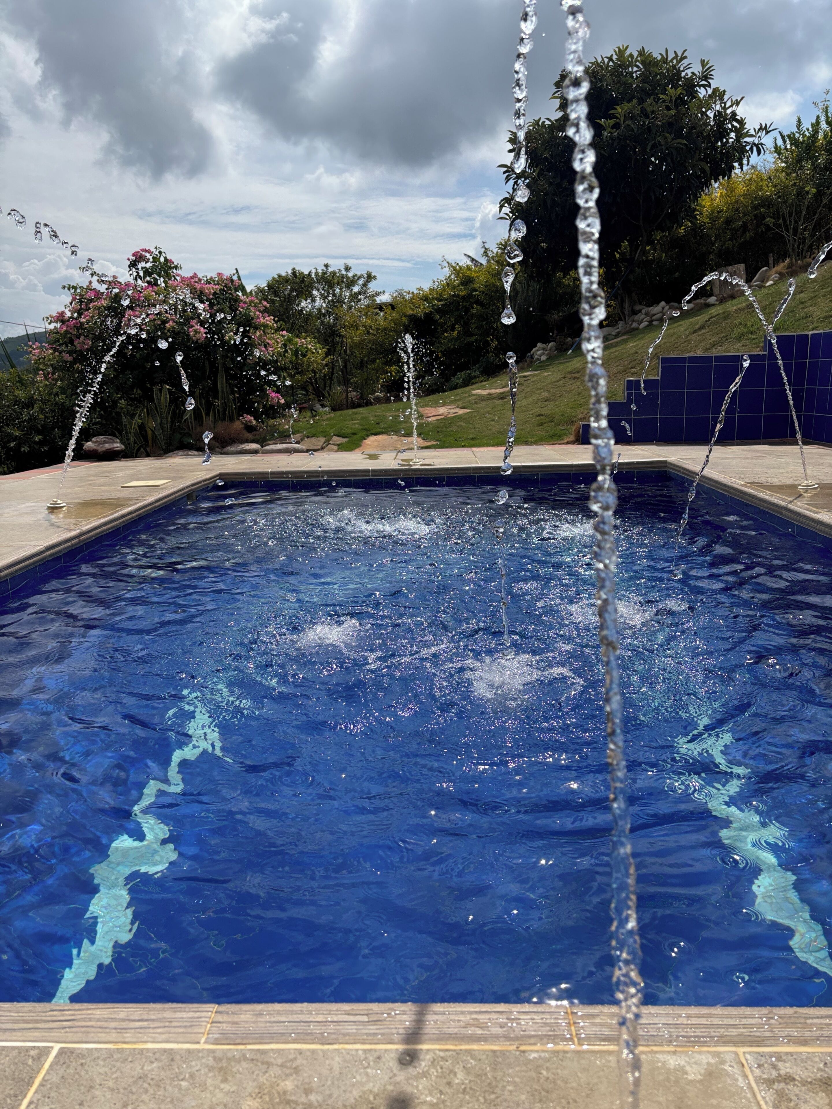 Una piscina con chorros de agua, rodeada por un solárium con baldosas y vegetación.