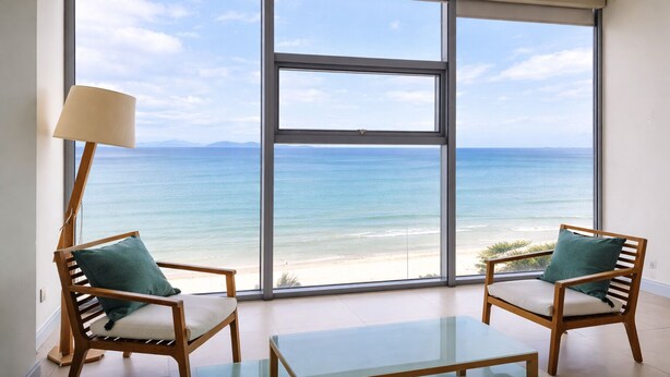 Una stanza con una grande finestra vista spiaggia, due poltrone di legno con cuscini, un tavolino in vetro e una lampada da terra.