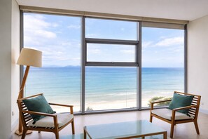 A room with a large window overlooking a beach, two wooden chairs with cushions, a glass table, and a floor lamp.