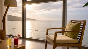 Una stanza con una grande finestra vista su uno specchio d'acqua, una sedia di legno con cuscino e due bicchieri su un tavolo.