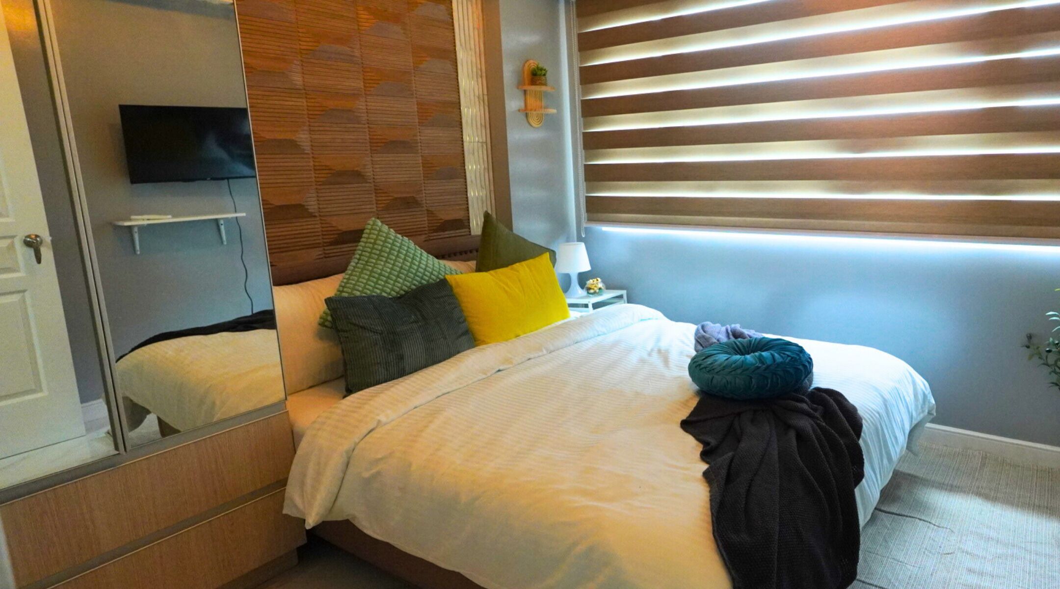 A hotel room with a bed, a TV, a bedside table with a lamp, and a window with blinds.