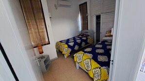 A room with two beds, a window with blinds, a lamp, and a door. - Cozy apartment in quiet area with fantastic sea view and swimming pool, (Riviere Noire)