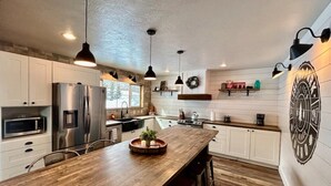 Cocina moderna con armarios blancos, electrodomésticos de acero inoxidable, una isla de madera, iluminación colgante y un emblema decorativo en la pared.