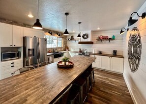A modern kitchen with white cabinets, stainless steel appliances, a wooden island, pendant lighting, and a decorative wall emblem. - TT CastIron Open layout, EV, close2Yellowstone, hotTub, AC (Island Park)