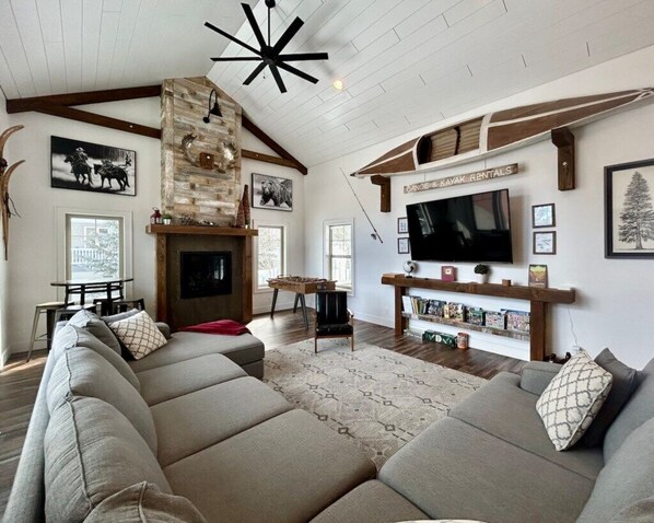 A spacious living room with a large sectional sofa, a fireplace, a mounted TV, and a wooden ceiling. - TTM Open layout, EV, close2Yellowstone, hotTub, AC (Island Park)
