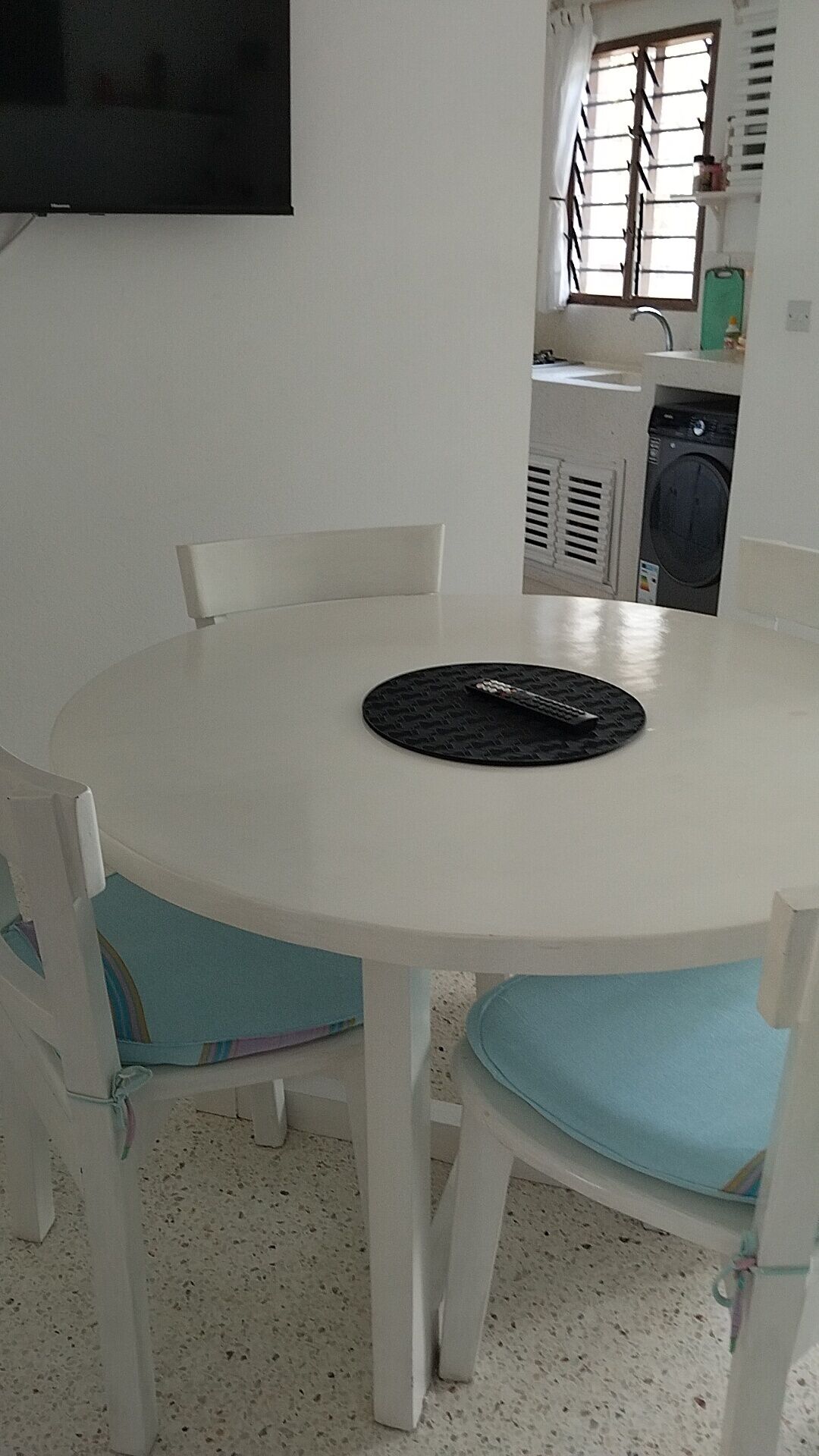 Una mesa redonda blanca con sillas acolchadas en azul, un mantel individual negro y un televisor montado en la pared.