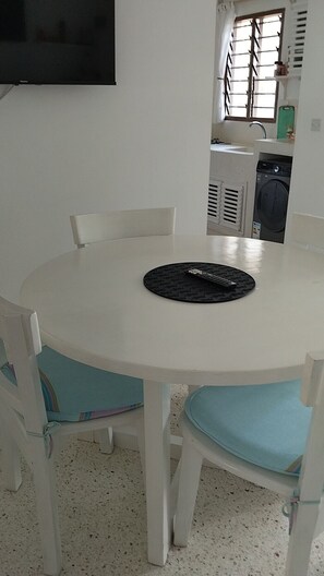 Una mesa redonda blanca con sillas acolchadas en azul, un mantel individual negro y un televisor montado en la pared.