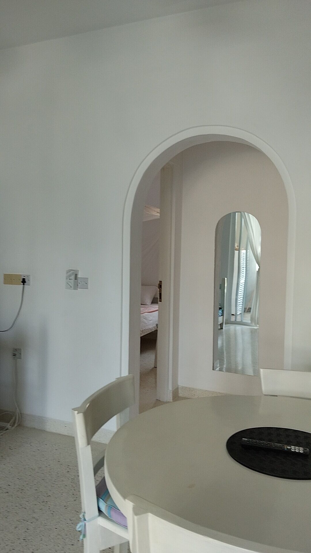 Ein Zimmer mit einem runden Tisch, einem Sessel, einem Bett im Hintergrund und einem Spiegel an der Wand.