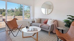 Ein modernes Wohnzimmer mit einer Couch, zwei Ledersesseln, einem Couchtisch, einem runden Spiegel und Blick auf ein Gebäude durch große Fenster.
