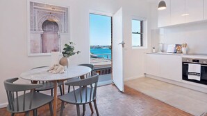 Eine moderne Küche mit weißen Schränken, eine Essecke mit einem runden Tisch und Stühlen sowie Blick auf das Meer durch eine offene Tür.
