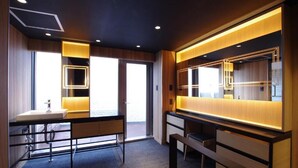 A modern hotel room with a large mirror, a desk with drawers, a bench, and a window providing natural light.