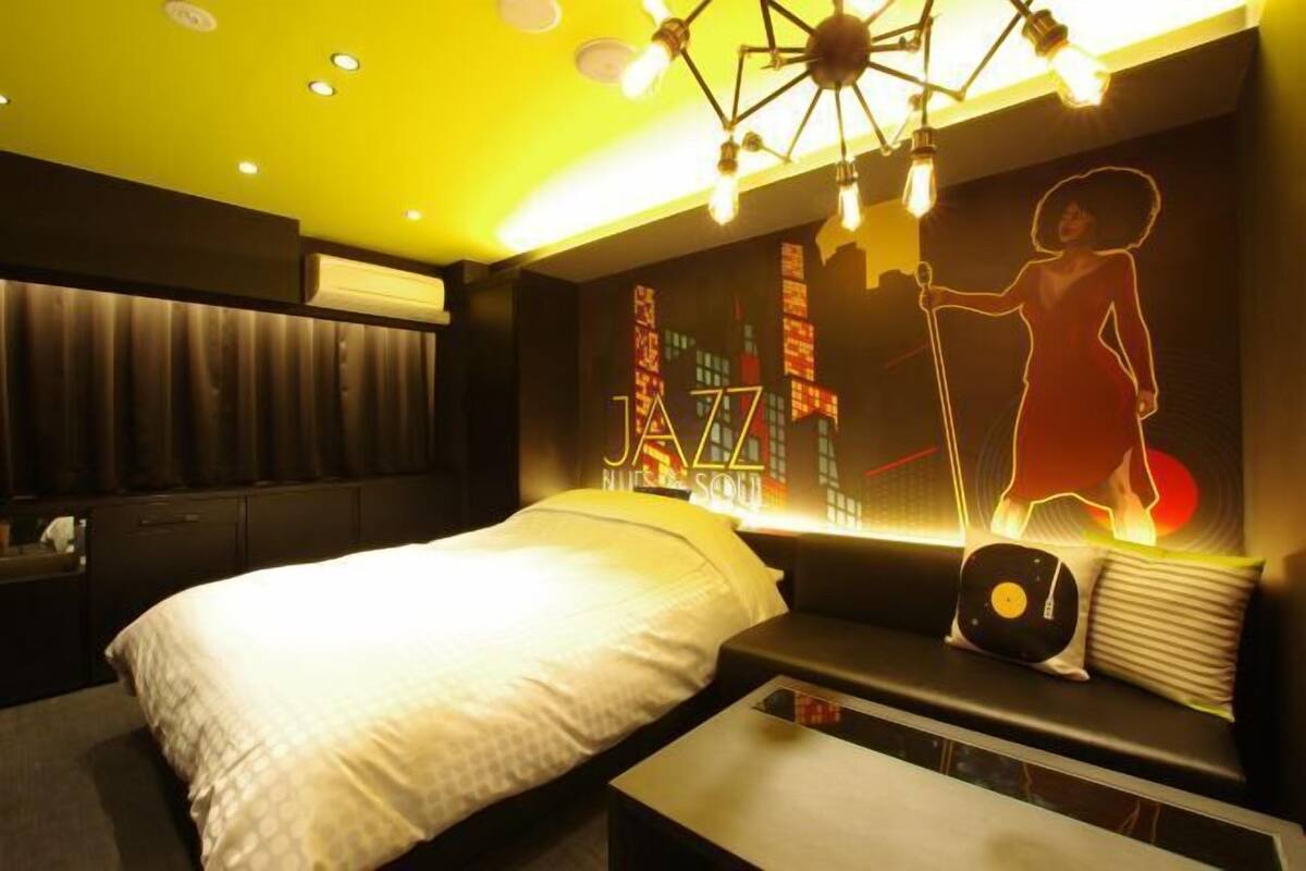 Une chambre d’hôtel moderne équipée d’un lit, d’un coin salon avec un tourne-disque et d’un mur orné d’une fresque représentant un paysage urbain.