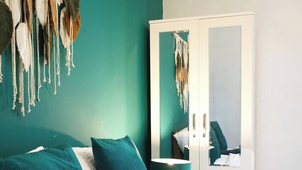 Un dormitorio con paredes verde azulado, una cama blanca, almohadas verde azulado y una manta blanca, además de un colgador de macramé en la pared.