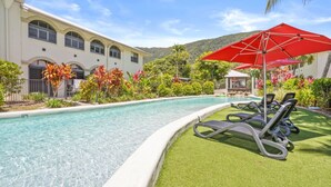 Ein Pool mit klarem Wasser, umgeben von einer gepflegten Rasenfläche, und ein Gebäude mit heller Fassade und Rundbogenfenstern.