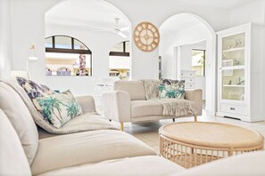 Un salon comprenant un canapé, un fauteuil et une table basse ronde, des murs blancs et une horloge en bois.