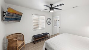Une chambre à coucher comprenant un lit, un téléviseur fixé au mur, un ventilateur de plafond, un banc et une porte avec des stores.