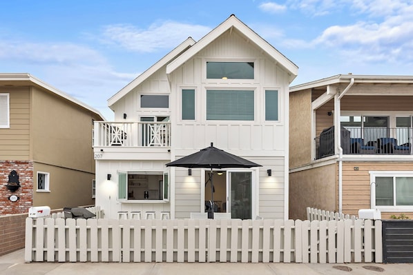 A two-story residential building with a balcony, a covered patio with an umbrella, and a white picket fence.