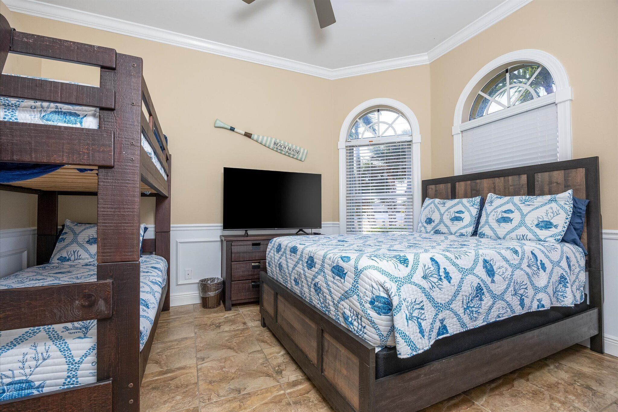A bedroom with bunk beds, a flat-screen TV, a bed with patterned bedding, and a window with blinds.