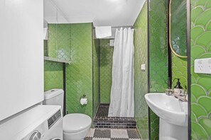 Casa de banho com paredes revestidas a azulejos verdes, uma sanita branca, um chuveiro com cortina branca e uma pia com espelho.
