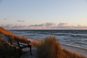 Eine Bank am Meer mit Wellen, grasbewachsenen Dünen und blauem Himmel.
