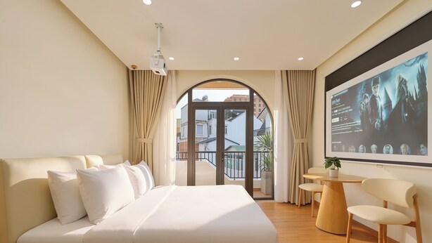 A hotel room with a large bed, a desk, and a TV screen displaying a movie scene. - Dalat Blanc Residence (Cam Ly - Da Lat)