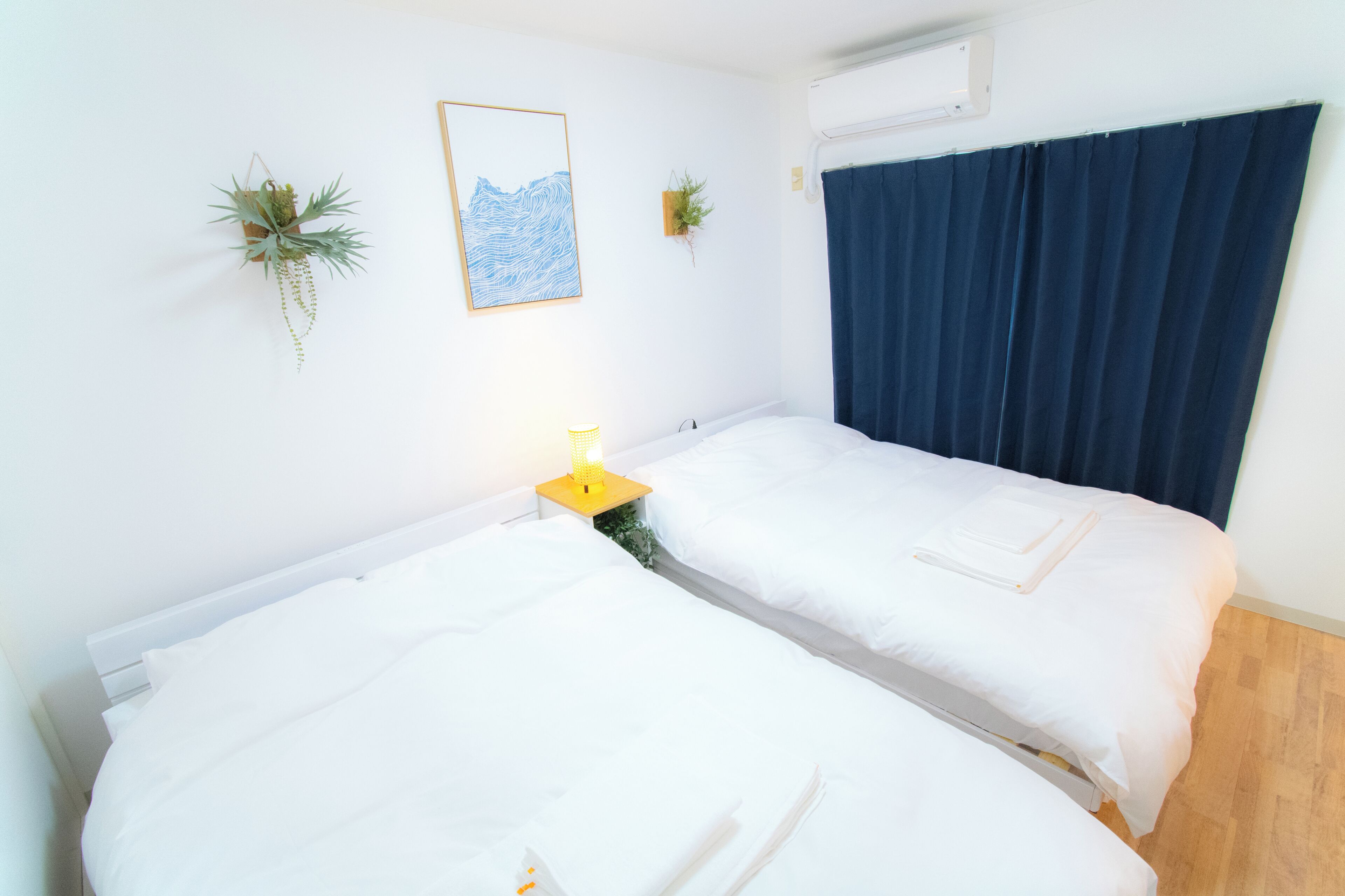 A room with two beds, a bedside table with a lamp, a wall-mounted air conditioner, and two potted plants.