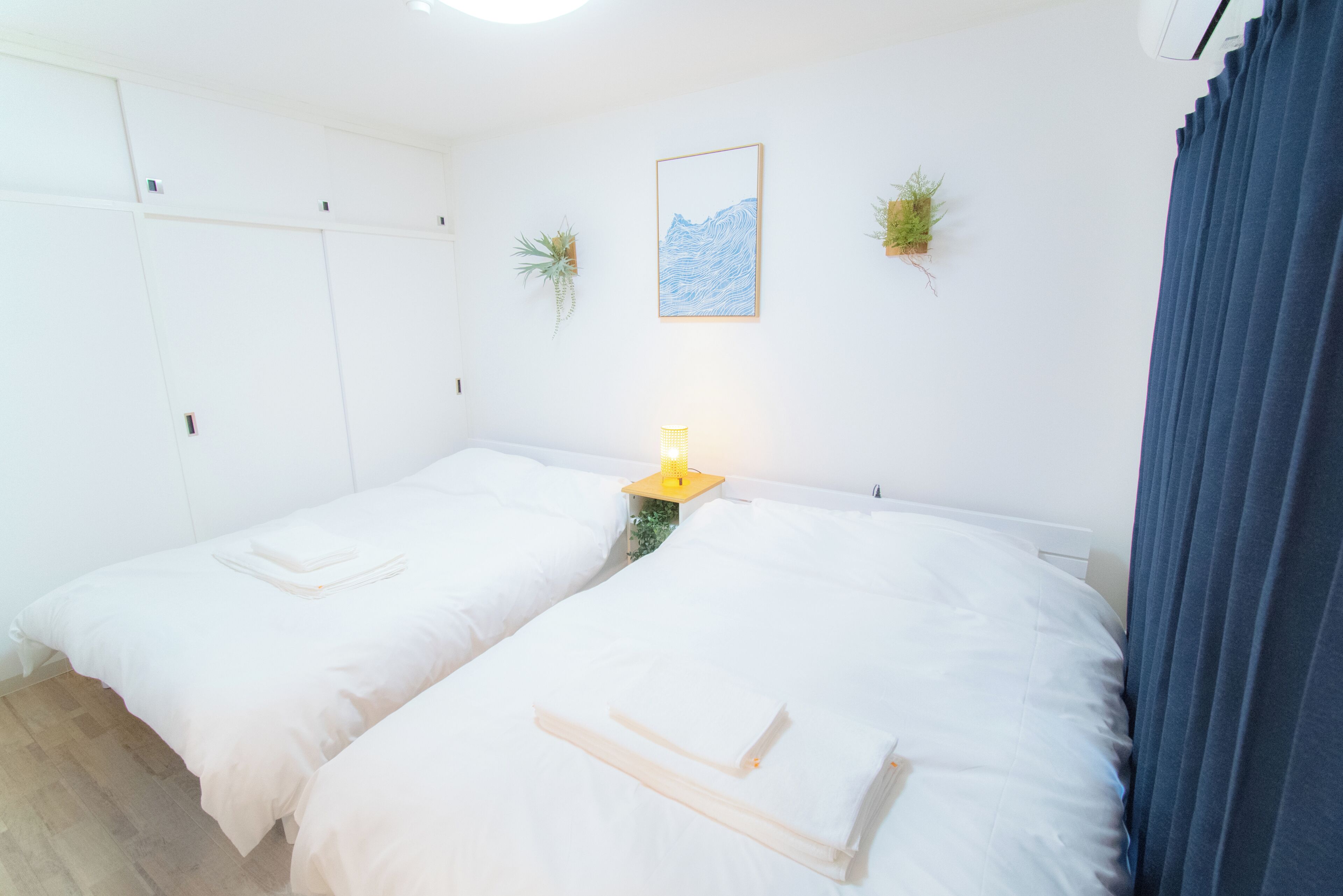 A room with two beds, a bedside table with a lamp, a framed picture on the wall, and two potted plants.