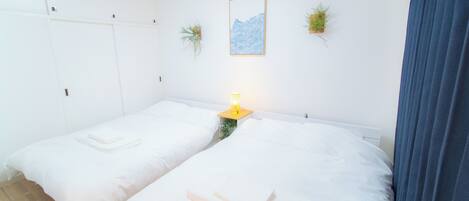 A room with two beds, a bedside table with a lamp, a framed picture on the wall, and two potted plants.