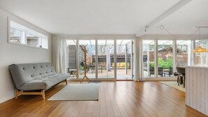 Una habitación amplia con sofá gris, suelo de madera, grandes puertas correderas de vidrio que dan a un espacio exterior, y una pequeña mesa con sillas.