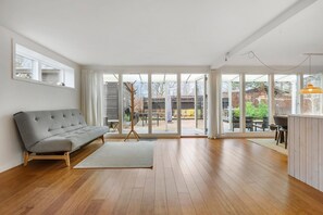 A spacious room with a grey sofa, wooden flooring, large sliding glass doors leading to an outdoor area, and a small table with chairs. - Lovely holiday home near beach and nature (Vejby)