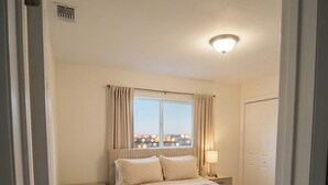 Una camera da letto con un letto, un comodino, una lampada, una finestra con tende e vista sulla città.