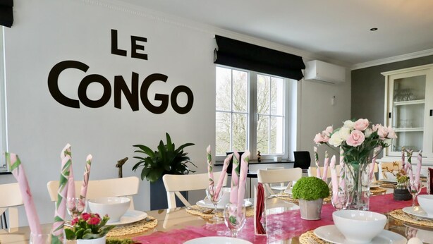 Un coin repas avec une table dressée pour le dîner, un mur sur lequel on peut lire le mot « CONGO », et une fenêtre donnant sur des arbres.