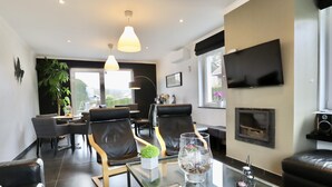 Un salon moderne avec une table basse en verre, des canapés en cuir, une cheminée, un téléviseur et un coin repas comprenant des chaises et une plante.