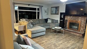 Ein Wohnzimmer mit einem Ecksofa, einem Kamin, einem Deckenventilator und einem großen Fenster.
