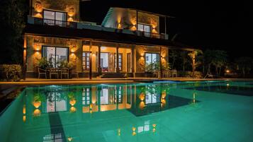 Un edificio de dos pisos con piscina frente a él, ventanas grandes e iluminación exterior.