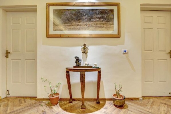 A room with a wooden floor, a framed painting on the wall, a small table with a statue and a vase, and two potted plants.