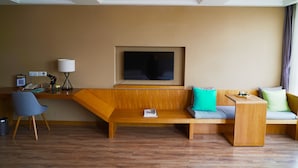 A modern hotel room with a wooden bench, a flat-screen TV mounted on the wall, a desk with a lamp, and a chair. - PengLai Mahota Town (Yantai)