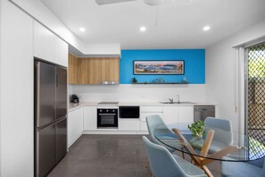 Eine moderne Küche mit Edelstahlgeräten, einer blauen Akzentwand und einem Essbereich mit einem Glastisch und blauen Stühlen.