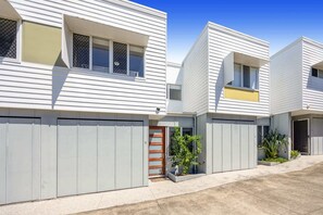 Ein modernes Wohngebäude mit symmetrischer Gestaltung, das einen zentralen Eingang mit einer Holztür, eine kleine Grünanlage mit Pflanzen und einen Betonweg zeigt.