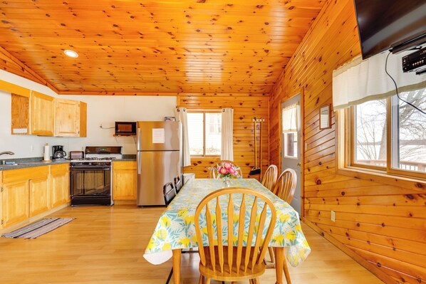 A cabin interior with wooden walls, a dining table with chairs, a kitchen area with appliances, and a television mounted on the wall. - Lake Access & Pier: Northwoods Fishing Cabin (Pearson)