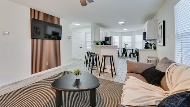 A modern living room with a ceiling fan, a TV mounted on a wooden panel, a dining area with stools, and a kitchen with a central island. - < 1 Mi to Historic McKinney: Modern Duplex w/ Yard (McKinney)